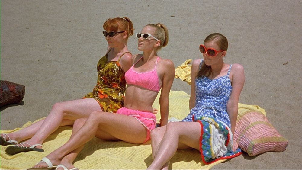 A trio of “teen” girls in swimsuits (yellow, pink, and blue) sitting on a yellow beach blanket on the beach