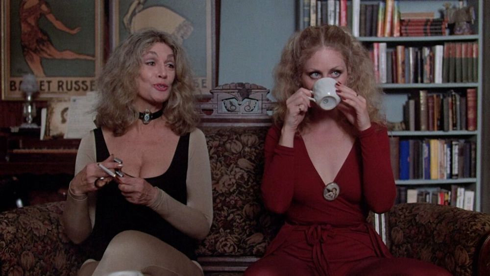 Two Blonde women sitting on a couch - one in tan and black on the left; one in red, sipping a white teacup, on the right