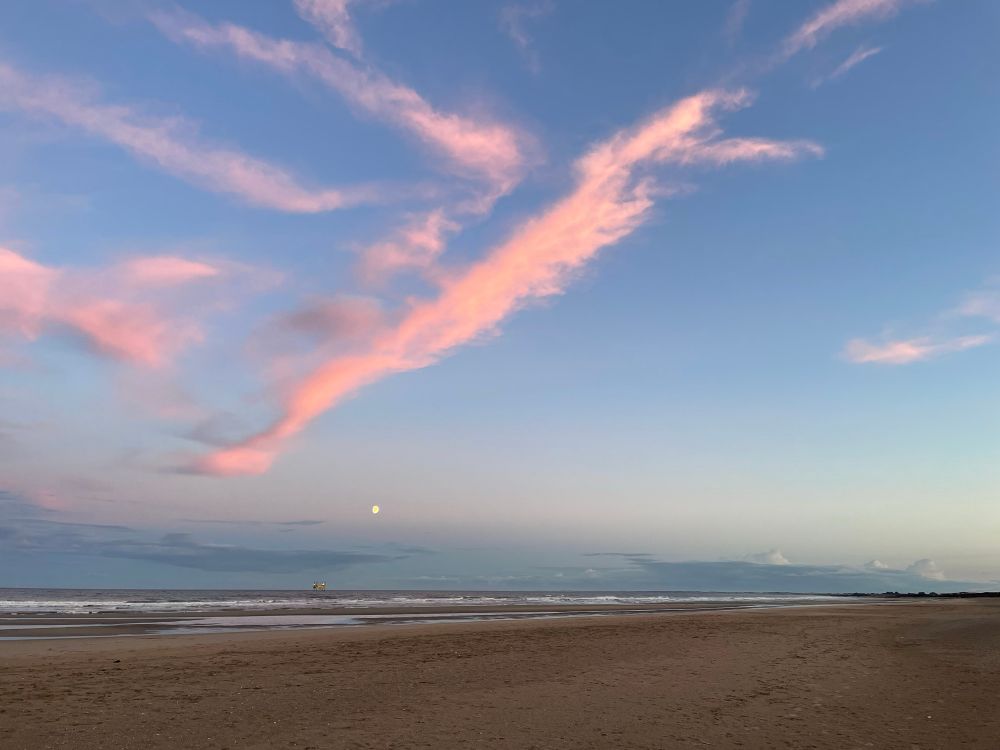 diagonal pinky streaks of cloud in a deepening blue sky. sandy beach along the bottom of the image and a glimpse of the low moon above the horizon