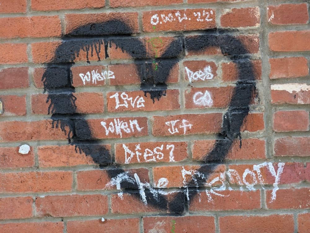 Graffiti, in white paint; surrounded by a black heart on a red brick wall. With 'The Memory" in white on top of it all