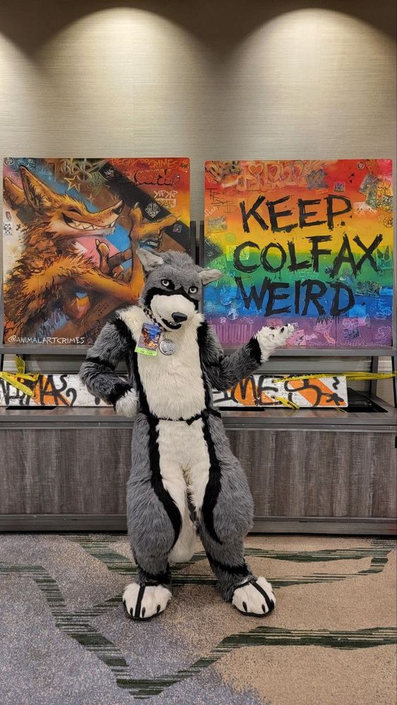 A Gray Wolf fursuiter with his arm outstretched, as if to present an animal art crimes art piece, with the message "keep Colfax weird" written on it. It also depicts the progress flag with the yop proudly ushering in the message. 