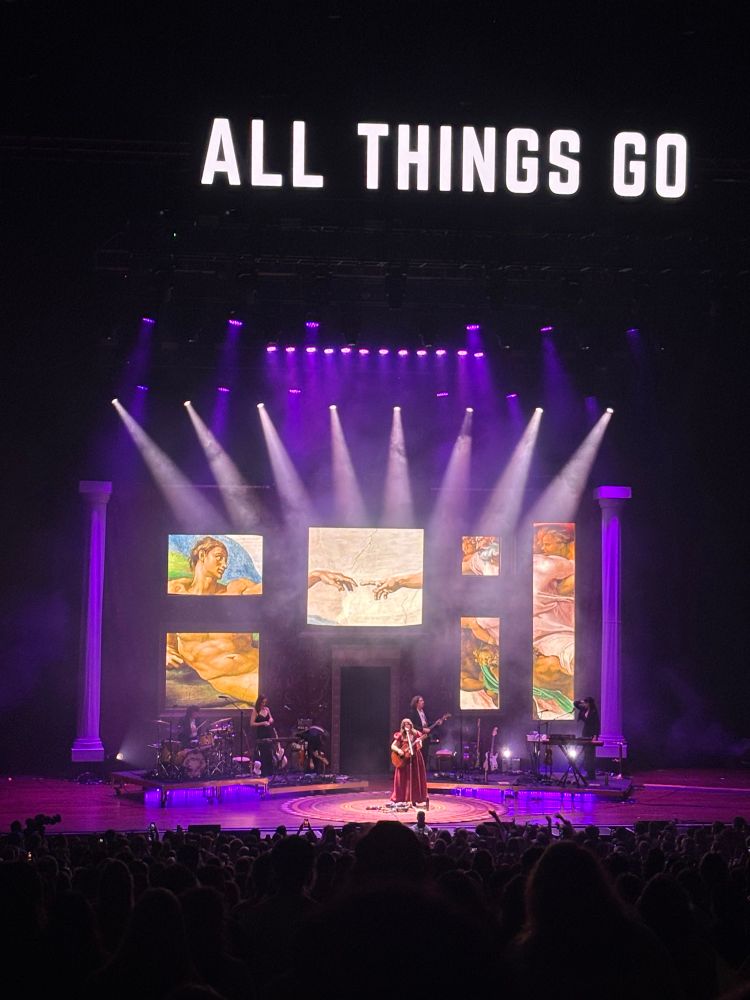 Photo of Lucy Dacus performing on All Things Go pavilion stage. The stage is lit in purple lighting with various art works including The Creation of Adam 