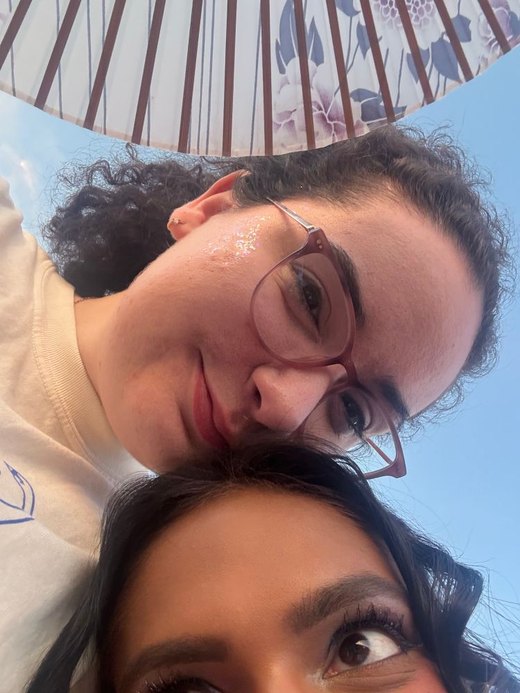 Close up photo of 2 young women smiling, one is holding a floral fan. 