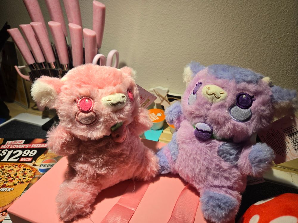 A pink cow plush with a strawberry on its collar, and a purple cow plush with a purple berry of some kind on its collar, sitting together on top of a pink gift box.