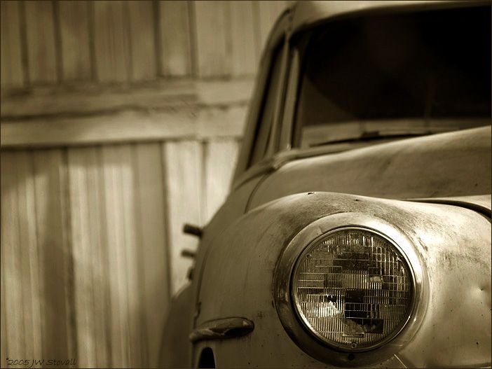sepia toned photo of the headlight and right front portion of an old car, in front of a wooden garage door.