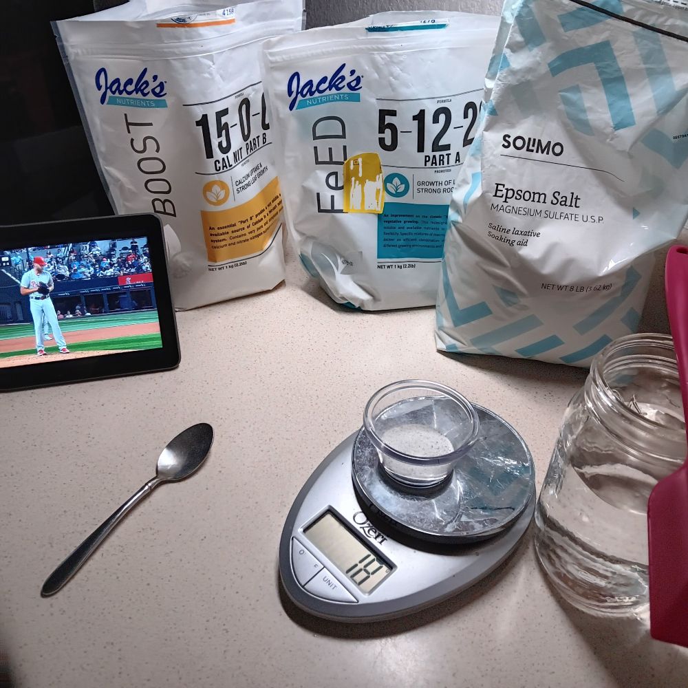 photo of a cream colored countertop with a digital scale on it weighing 18grams, two different bags of Jack's nutrients, a bag of epsom salts, a metal teaspoon, a glass mason jar with r/o water in it...and red silicon spatula laying on top of it. To the far left is a tablet with the game on....Wheeler,the pitcher, is on the mound.