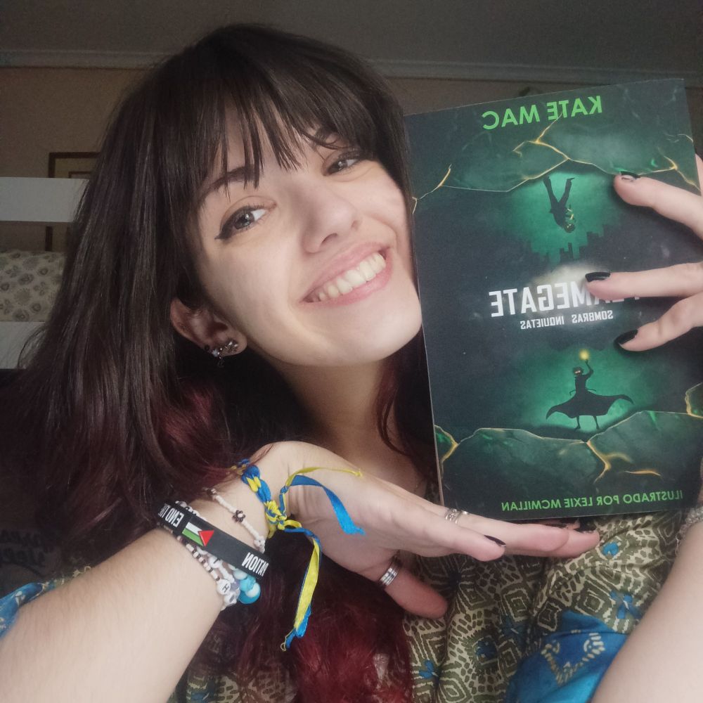 Picture of a white person holding up a paperback book. They're smiling and have dark hair, and they're wearing rings, black nail polish, and bracelets - one with the Palestinian flag, and one with the Ukrainian flag colours. 

The book they're holding up is the Spanish translation of "Flamegate. Restless Shadows", by Kate Mac.