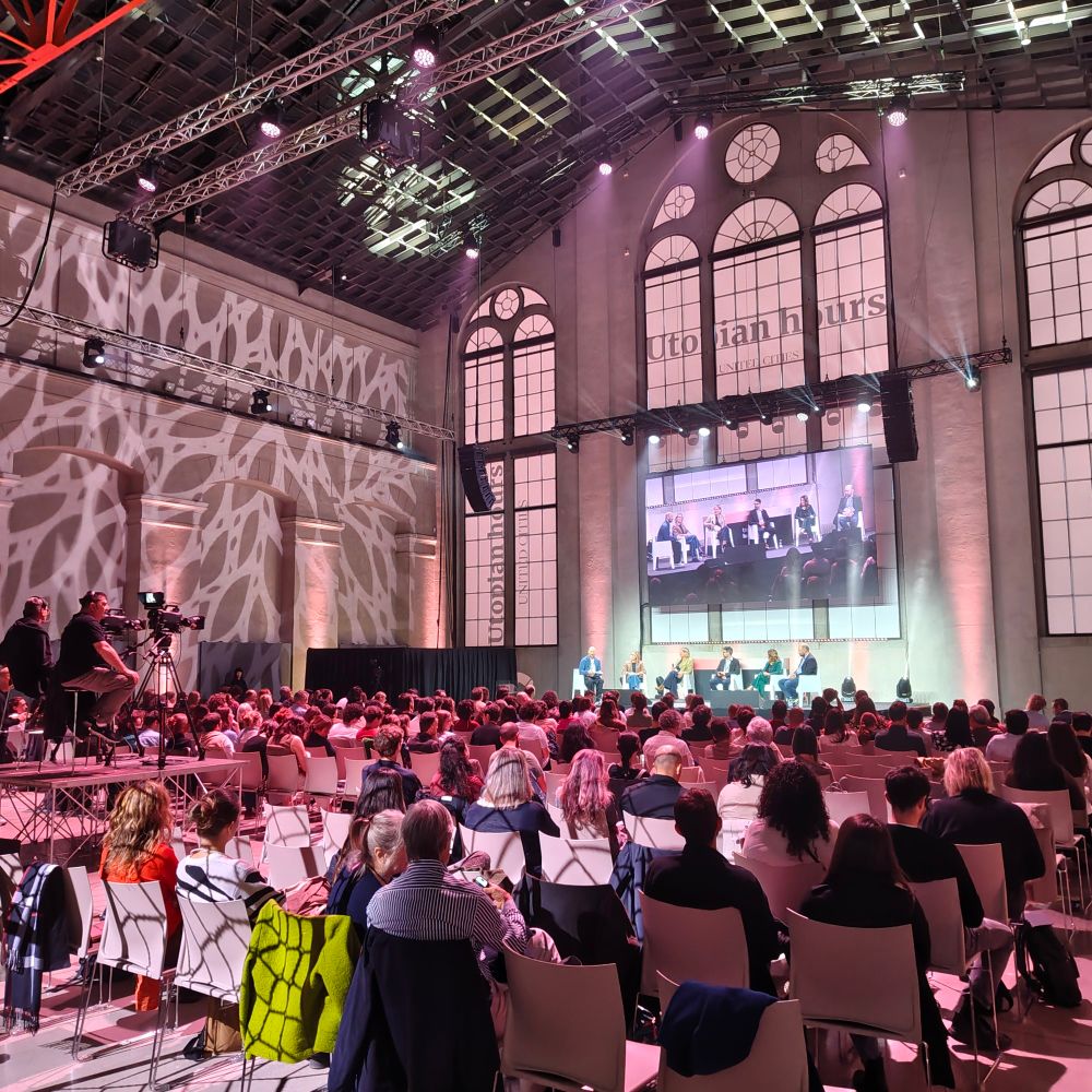 A large auditorium in the nuvola Centrale, Turin. The fire is from behind a seated crowd, facing a stage with a large screen over it.
