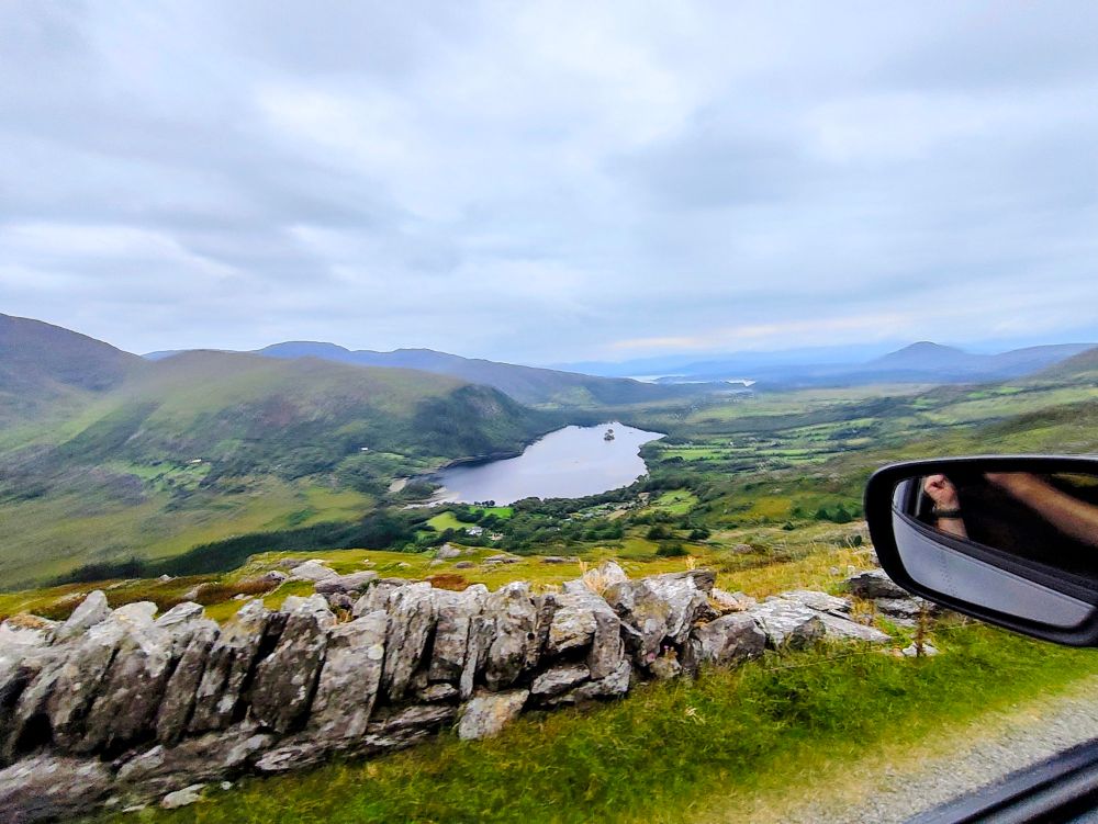A view out a car window, of a lake at the bottom of a valley, with a stone wall in the foreground.