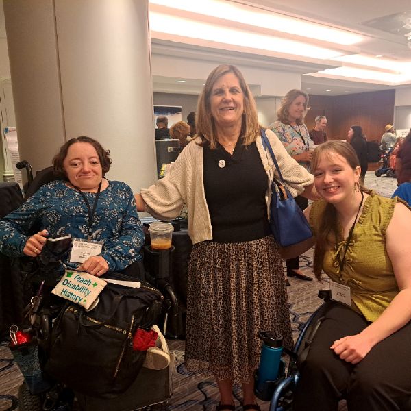 Three women at the 2025 NCIL Conference. Two women who use wheelchairs flank a standing woman with long blonde hair. One wheelchair user displays a "Teach Disability History" sign. All wear conference badges and are smiling