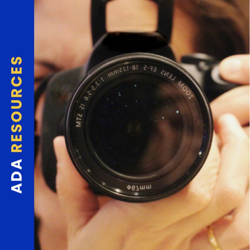A close-up image of a person holding a DSLR camera with their finger on the shutter button. The camera lens is centered in the frame, reflecting small lights. Along the left side, a vertical blue banner reads “ADA RESOURCES” in bold yellow and white text.
