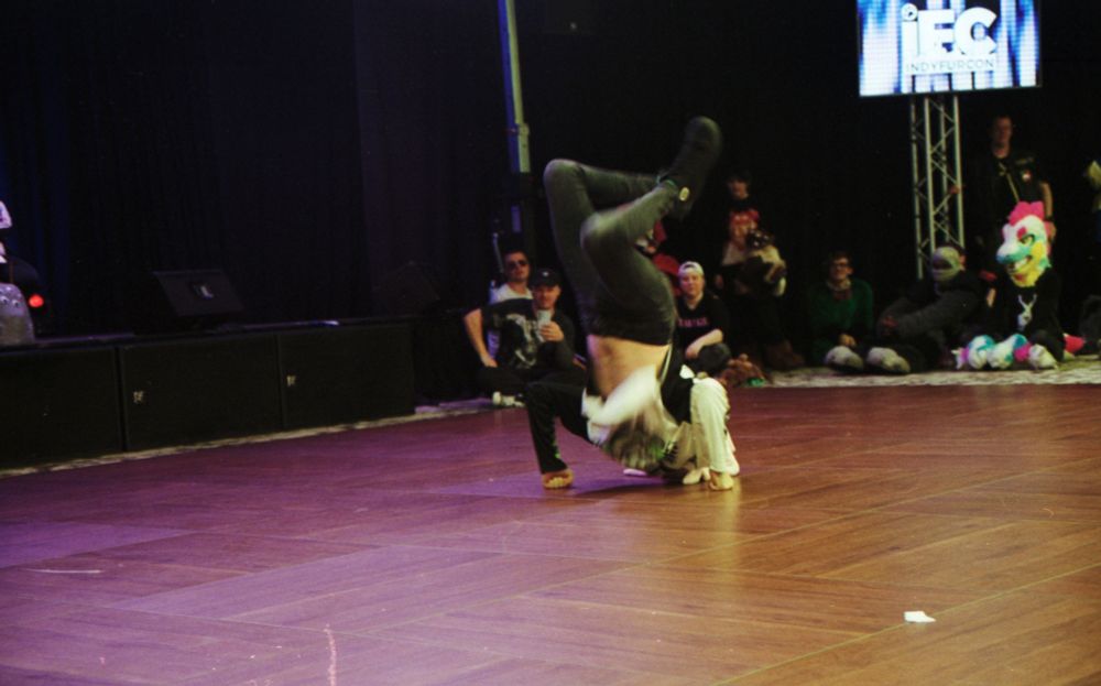 Photo of the same dancer, on the floor doing a move, arms bent, pushing up and legs kicked over the head and behind.