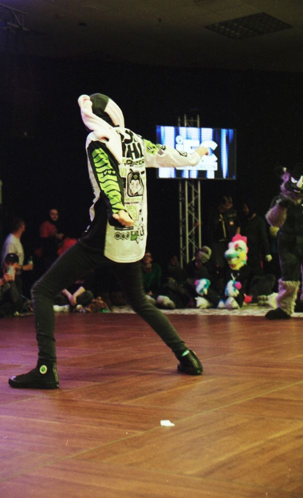 Photo of a dancer in streetwear and a hat with long floppy pink ears standing in a wide pose, knees slightly bent, arms spread behind.