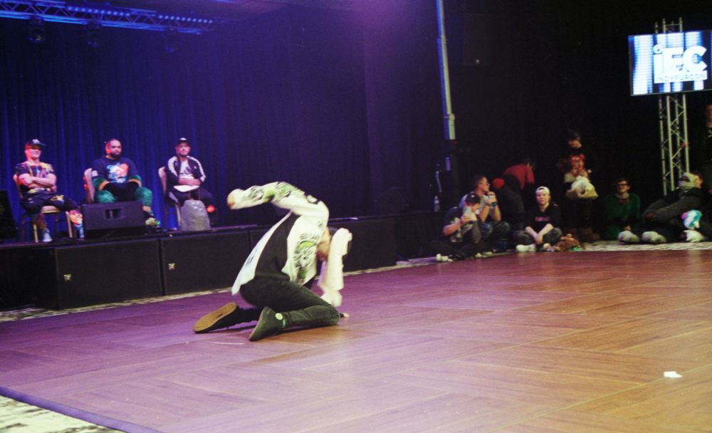 Photo of a dancer kneeling on the ground, bent forward, one arm raised behind their back. The ears of the dancer's hat obscure their face.