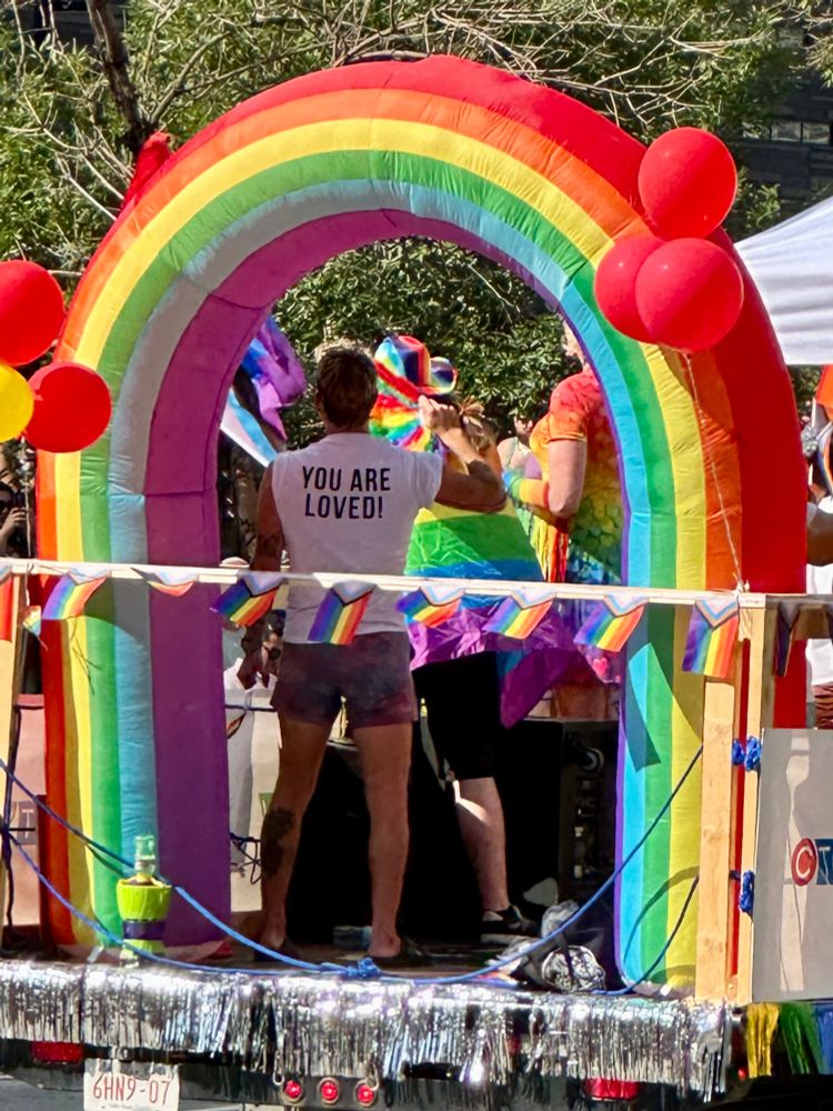 A rainbow arch with someone standing under it wearing a T-shirt that says “You are loved!” 