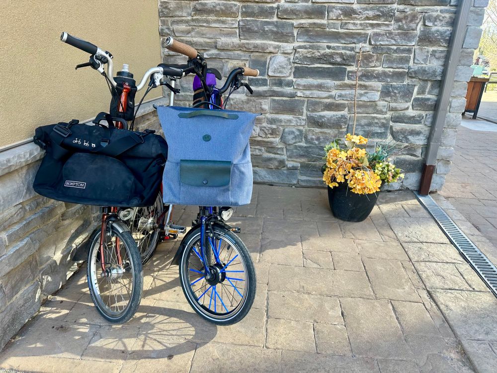 A photo of two folded bicycles tucked together next to a flower pot. 