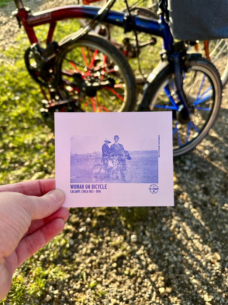 A printed postcard of a woman on a bicycle held in front of a folded bicycle. 