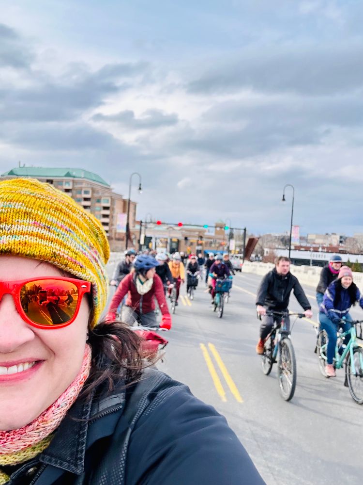 A selfie of a woman in red sunglasses with a yellow toque on, and a lot of bicycles are riding along behind her. 