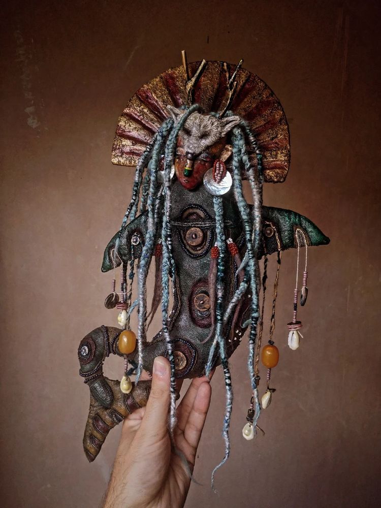Photo of a hand holding a textile art doll in the shape of a whale with human face and long dreaded hair, with beading and artic fox mask headdress and driftwood antlers