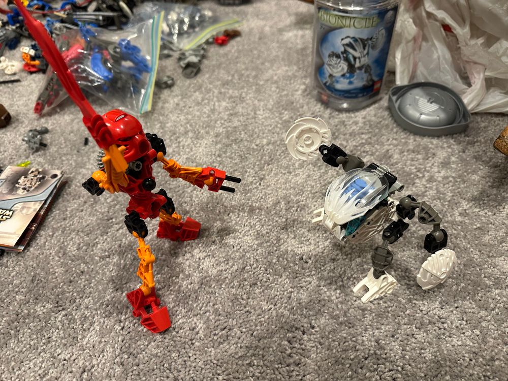 The original Tahu BIONICLE toy and a Kohrak Bohrok, assembled in a field of loose BIONICLE parts on a carpeted floor. They’re in battle poses. 