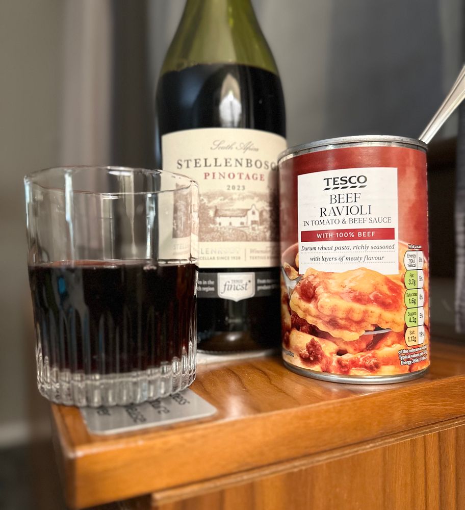 A can of Tesco beef ravioli next to a bottle and glass of “Tesco’s Finest” wine, sitting on a wood-panel radiator enclosure that’s probably older than I am. It’s not a fork sticking out of the can this time, it’s a spoon from the hotel tea set. The wine is honestly not terrible. 