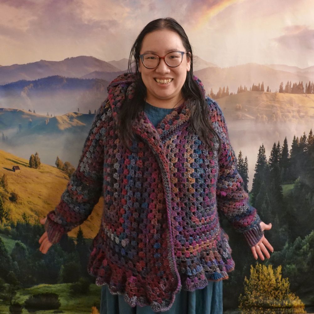 me, smiling in front of a tapestry of purple and golden and forest green hills, wearing a colorful granny hexagon cardigan in dark pinks, purples, and blues, my arms outstretched beside me