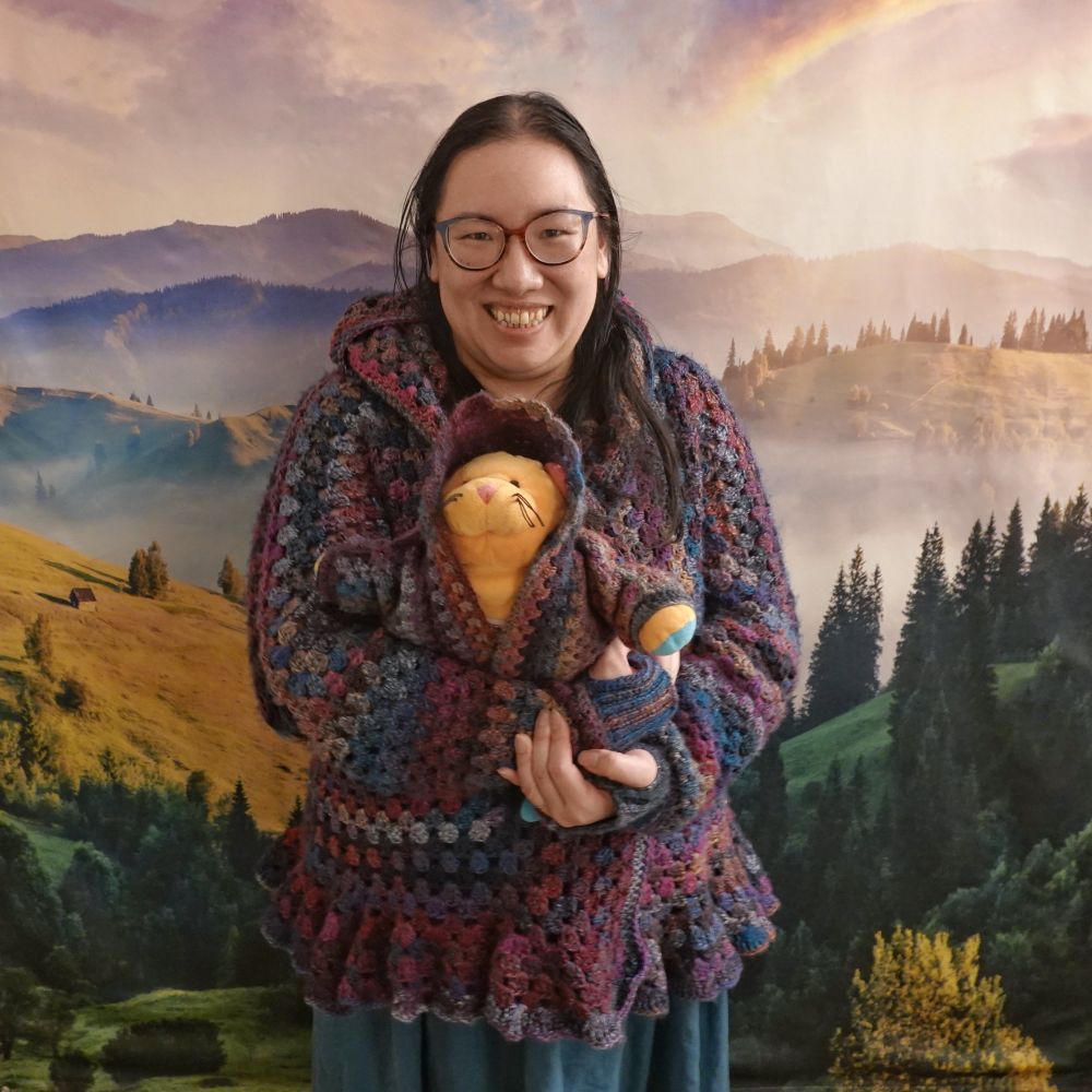 me, smiling in front of a tapestry of purple and golden and forest green hills, wearing a colorful granny hexagon cardigan in dark pinks, purples, and blues, holding a neon orange stuffed cat wearing a matching cardigan
