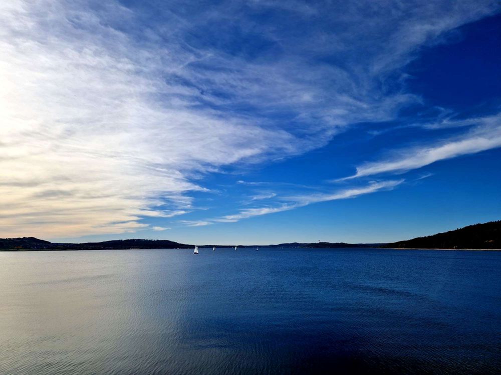 Ein blauer See, dessen Oberfläche durch den Wind leicht gekräuselt wird. In der rechten Bildhälfte zieht leichte Bewölkung auf, und in der Mitte sieht man winzige Segelboote mit weißen Segeln.