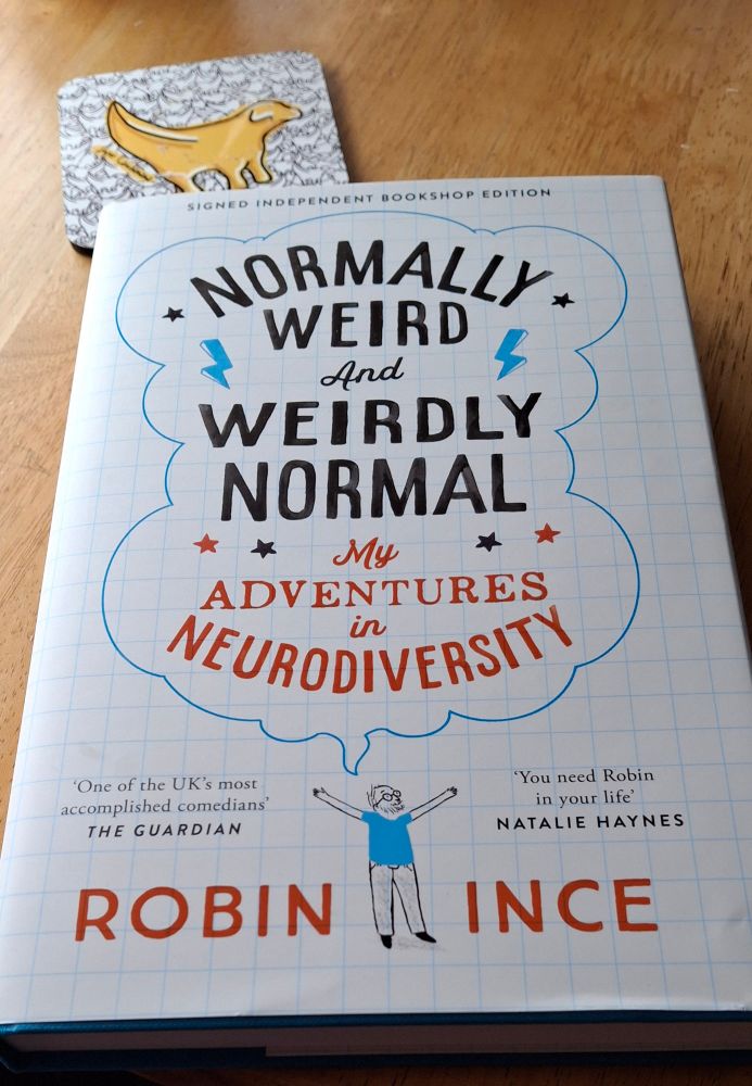 A book with a joyous figure of a mam in blue with words normally weird weirdly normally as a title. There is also a lambanana coaster.