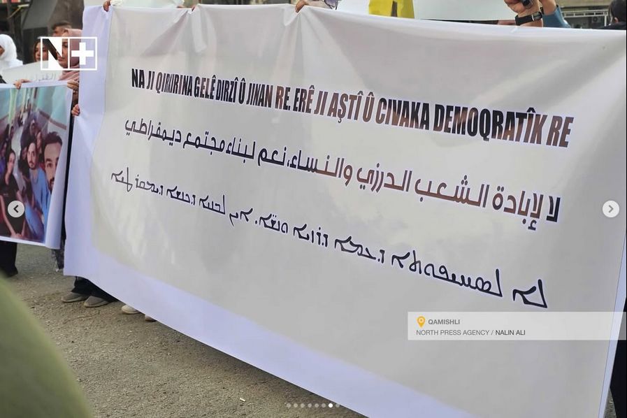 Auf einem großen weißen Transparent in schwarz auf weiß: "Nein zum Völkermord an den Drusen und ihren Frauen. Ja zum Frieden und einer demokratischen Gesellschaft", einmal auf Kurmandschi, Arabisch, und Assyrisch.

Oben links Logo von "N+", unten rechts "Qamishli North Press Agency / Nalin Ali".