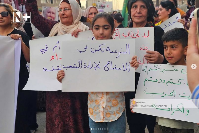 Eine Menge von Frauen und Kindern. Vorne hält ein Kind ein Schild mit dem Text "الشرعية تكمن في الاستماع لإرادة الشعب" bzw. "Legitimität liegt darin, auf den Willen des Volkes zu hören"

Oben links Logo von "N+", unten rechts "Qamishli North Press Agency / Nalin Ali".