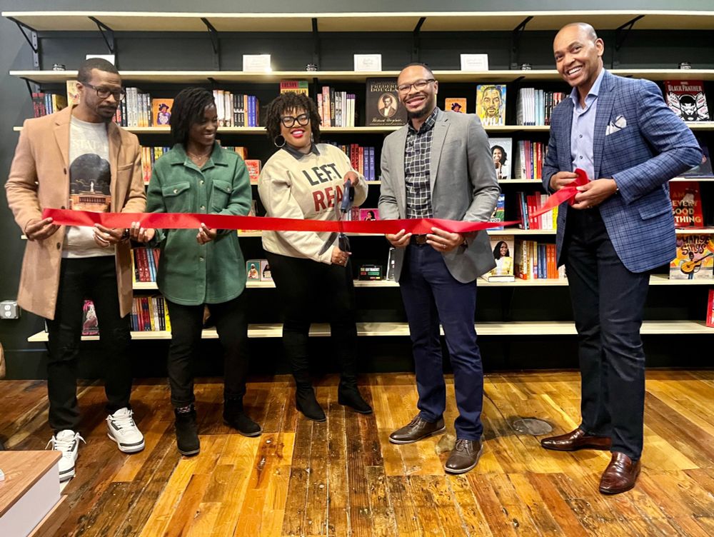 A photo of four people in a bookstore about to cut a red ribbon with a giant pair of scissors
