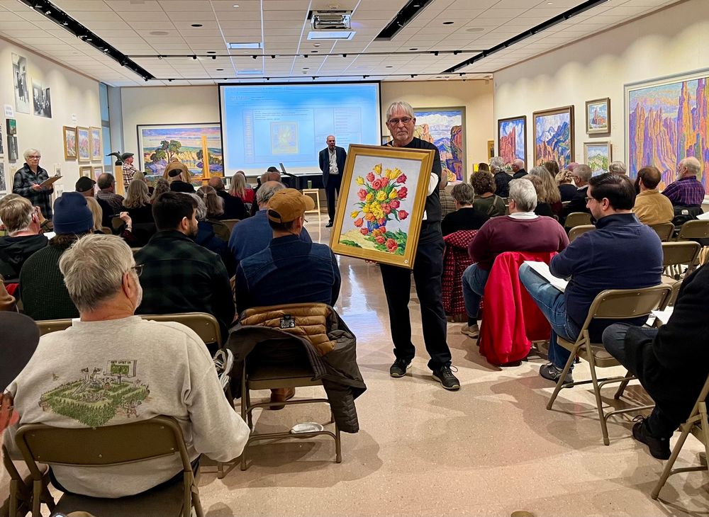 A photo taken from the back of the first gallery space at the Birger Sandzén Memorial Gallery. Rows of people face the auctioneer, who stands in front of a screen losing the current auction item. In the middle of the aisle, one of the auction works holds a painting of tulips in a vase by Birger Sandzén, a Swedish-American post-impressionist painter. 