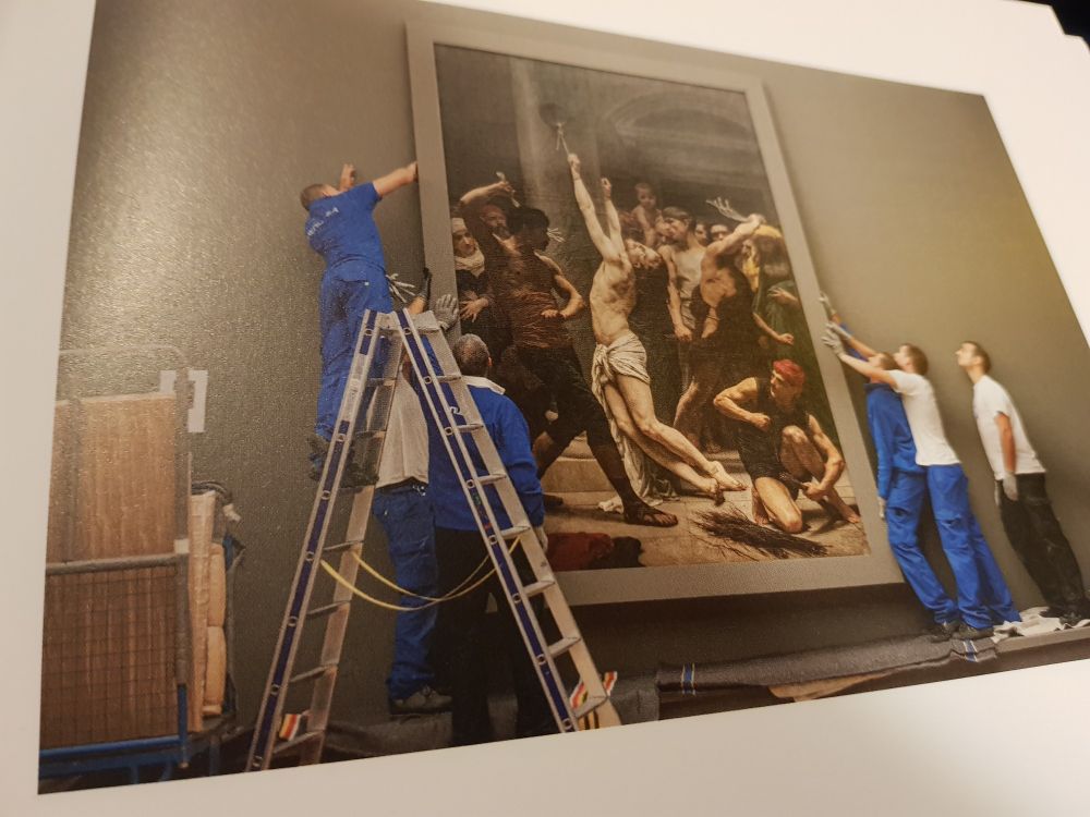 Six hommes, en bleu de travail accrochent ou décrochent une toile monumentale. Ce qui est peint et leur propre attitude forment comme un echo.