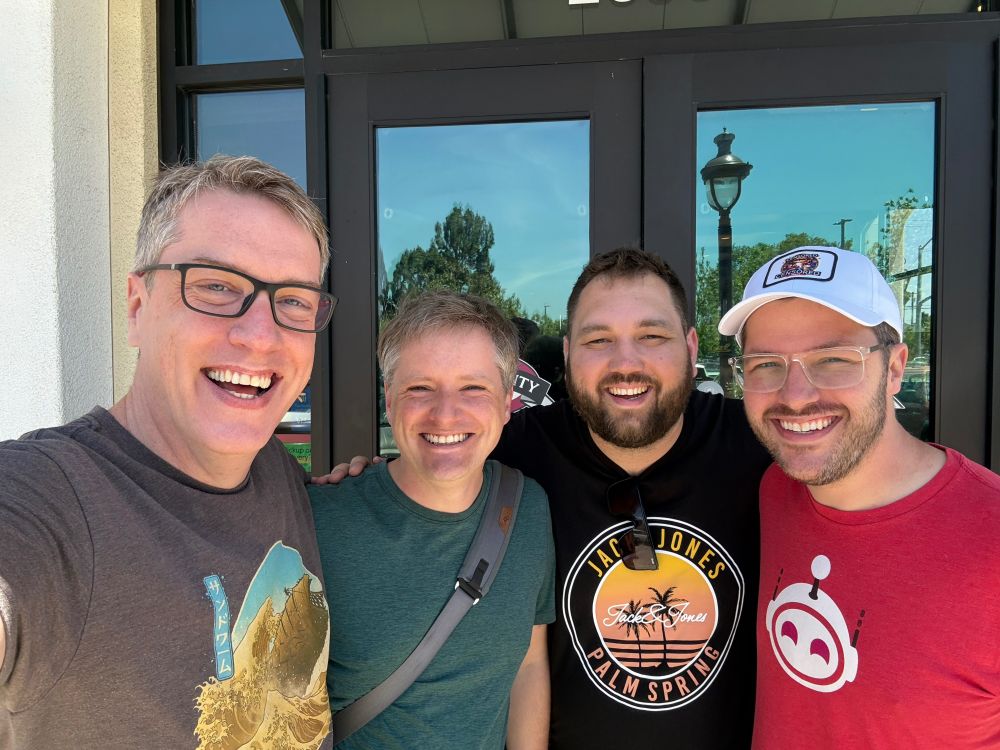 Four men smiling at the camera. One of them is wearing an Apollo T-shirt, because Apollo is still alive, damn it.
