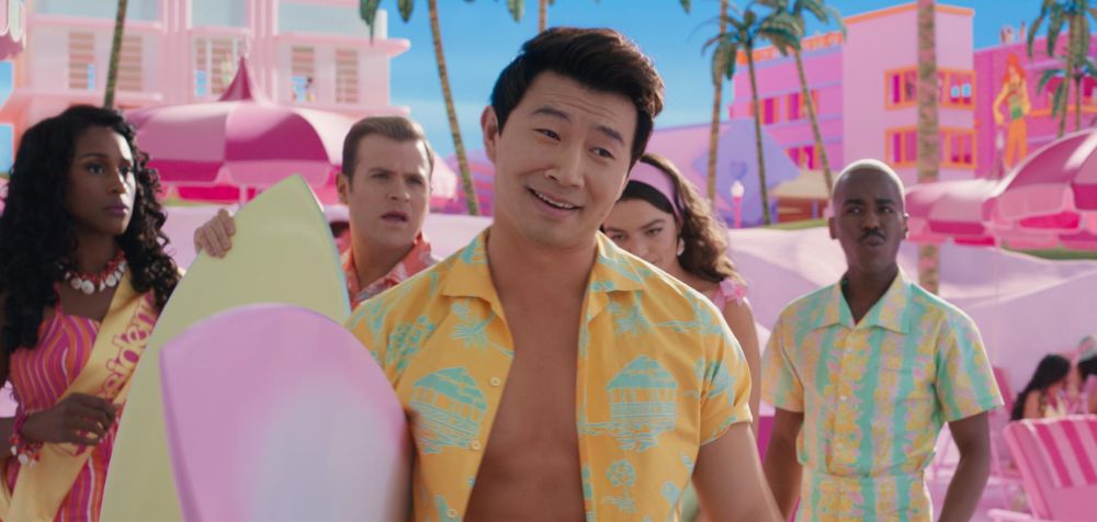 begin image ID: an image from the barbie movie. simu liu is in an unbuttoned yellow hawaiian shirt standing with several others in beach attire in front of pink beach houses. end image ID.