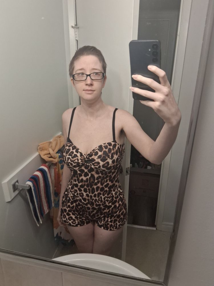 A white woman with short dark brown hair and glasses wearing a leopard print romper. She stands in the bathroom mirror.