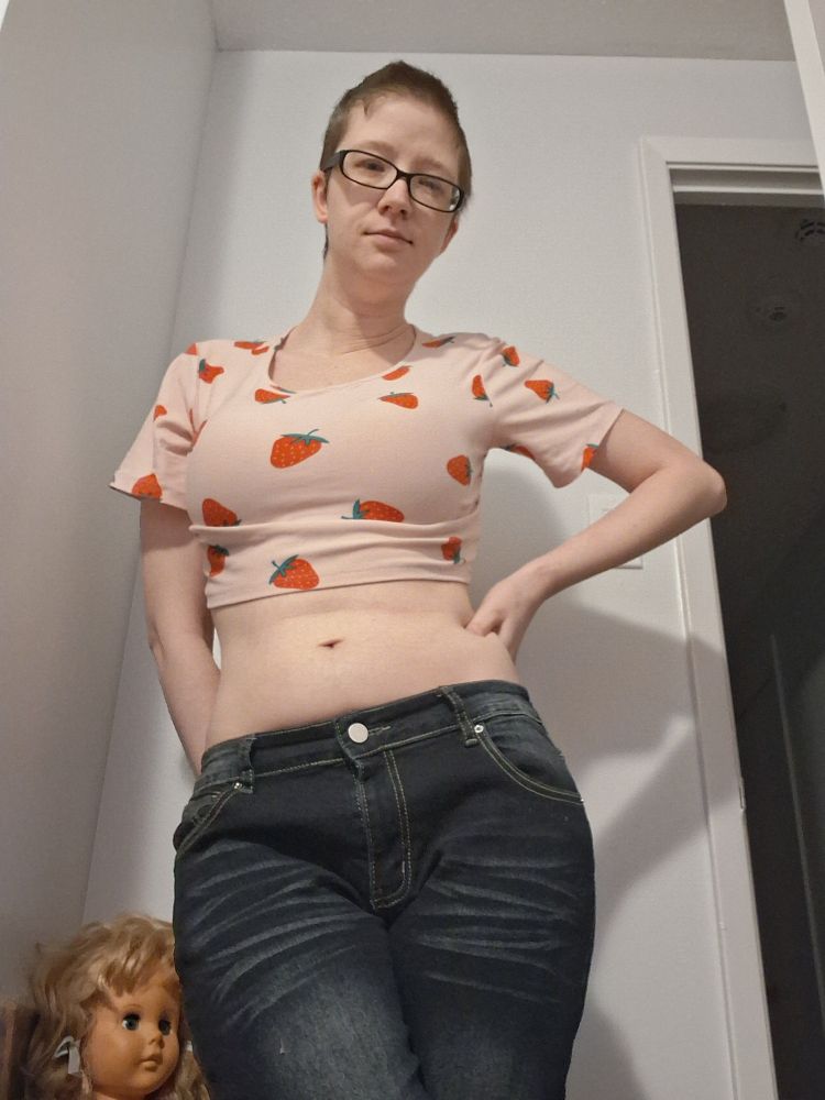 White woman with short dark brown hair and glasses wearing a pink crop top with strawberries on it and dark blue jeans. Posed with her hand on her waist.