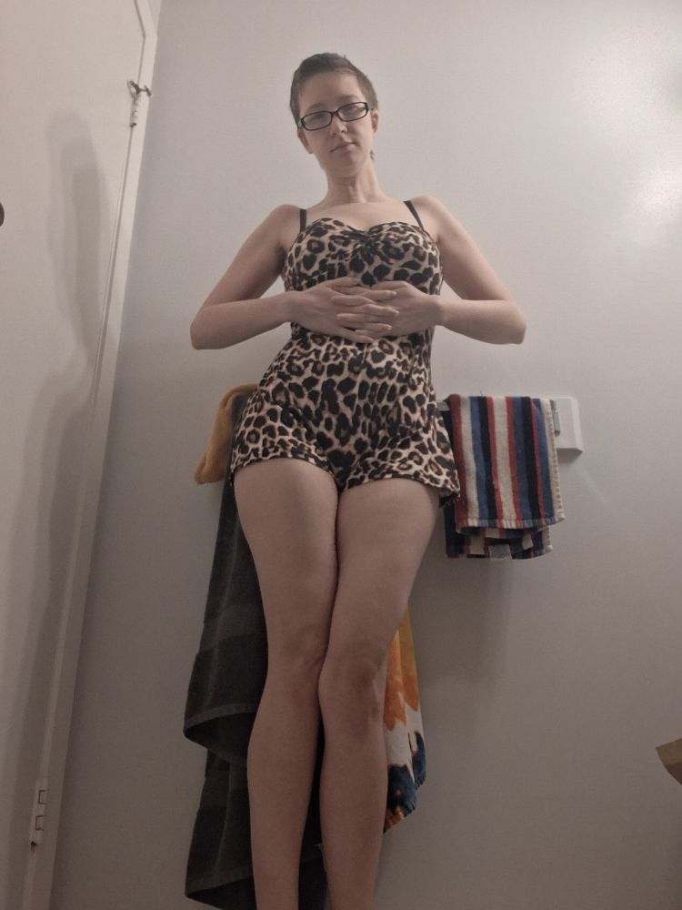 A white woman with short dark brown hair and glasses wearing a leopard print romper. Posed with her fingers interlocked across her abdomen.