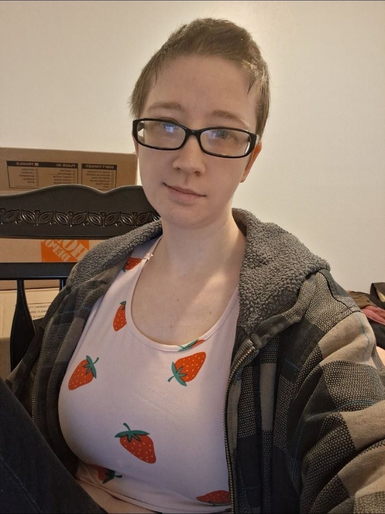 White woman with short dark brown hair and glasses wearing a pink crop top with strawberries on it, grey plaid hoodie and dark blue jeans. She's sitting in a chair, with her knee up. 