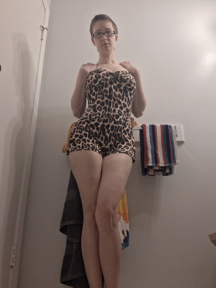 A white woman with short dark brown hair and glasses wearing a leopard print romper. Posed with her hands up touching her shoulders, one leg slightly crossed over the other one.