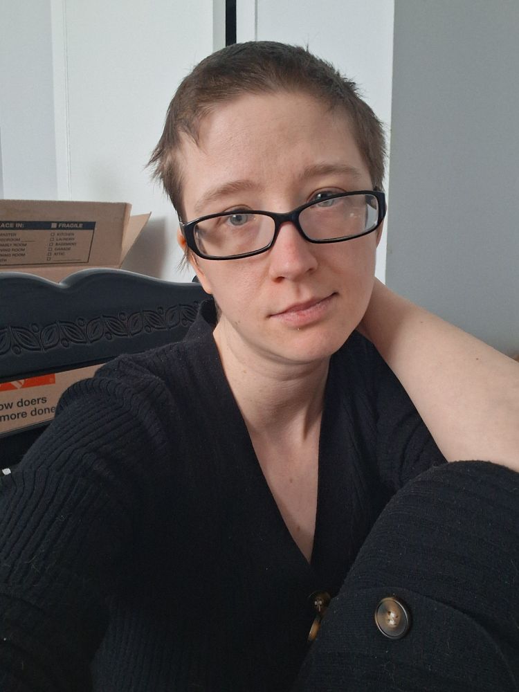 White woman with short dark brown hair and glasses wearing a black dress with faux buttons down the middle. Shes sitting in a chair with her knee up and shes holding the side of her neck.