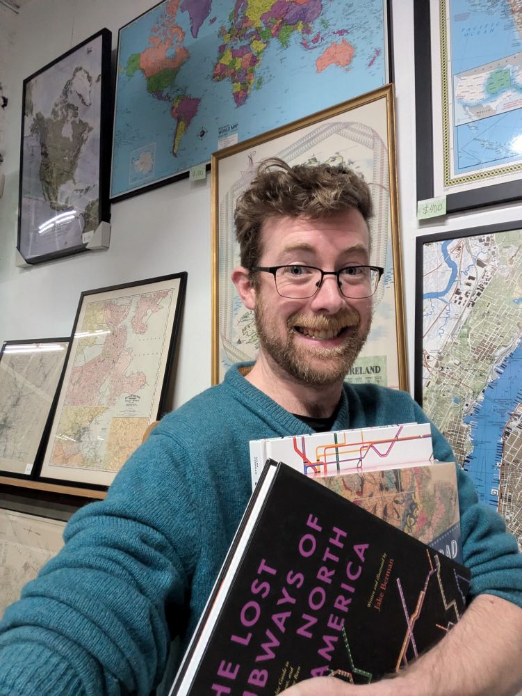 A nerd in a teal sweater holding a stack of books about railroads in front of a wall of framed maps.