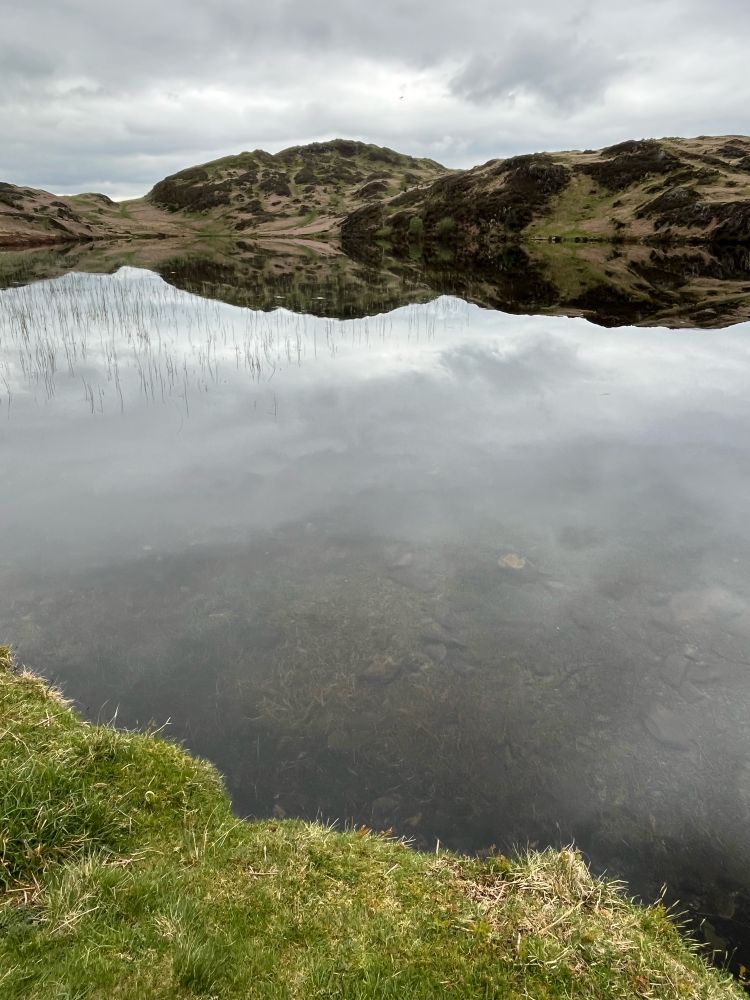 A brooding tarn. Still water, ragged diagonal of foreground grassy shore. Grey sky reflected in the water, dark fading to light meeting a reflected hilly horizon forming a lumpen horizontal band. Grey cloudy sky with soft white topped cloud. Not a waterfall in sight.