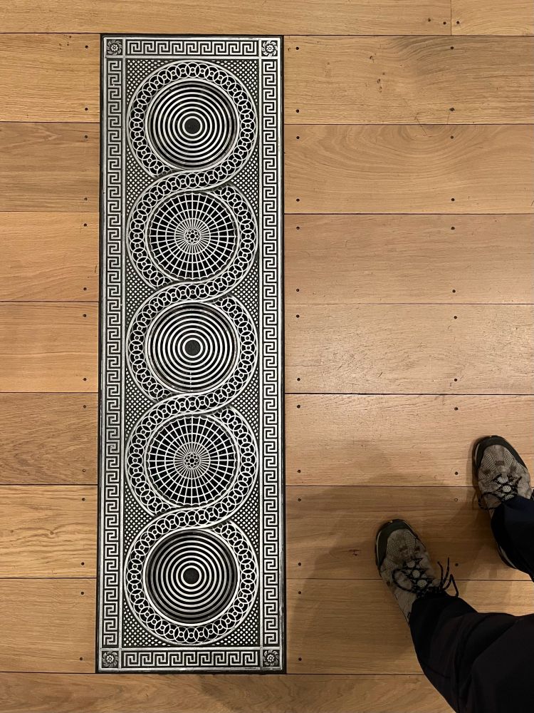When grilles were grilles. A wooden floor with boards running horizontally into which is set a metal ventilation grille. My feet are bottom right just for scale. The grille is a rectangular metal plate pierced to allow the passage of air from below the floor. The intricacy of the piercing is quite remarkable. A rectangular border frames five circles themselves bordered by a sinuous border linking (and separating) them. The outer two circles and the central circle are filled with diminishing concentric circles, the remaining two circles are perforated with a pattern of lines radiating from the centre. The work is as good as a silversmith’s and quite beautiful. Most people were looking at the Monets on the walls. 