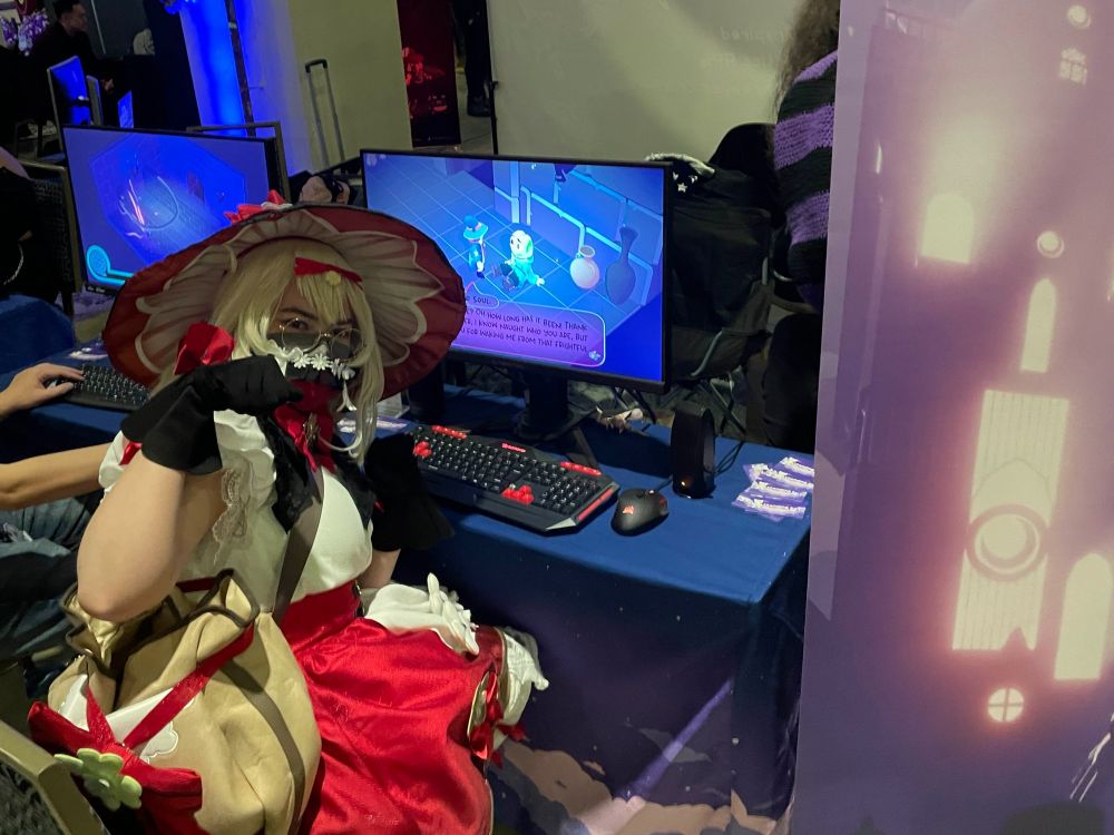 A cosplayer strikes a pose while sitting at the Nekomancer of Nowhere booth! The captioner does not know which character, but the outfit is red, white, and giving cottage-core. In-game, they've left the nekomancer talking to a frog ghost who kicks its legs while sitting on the floor.