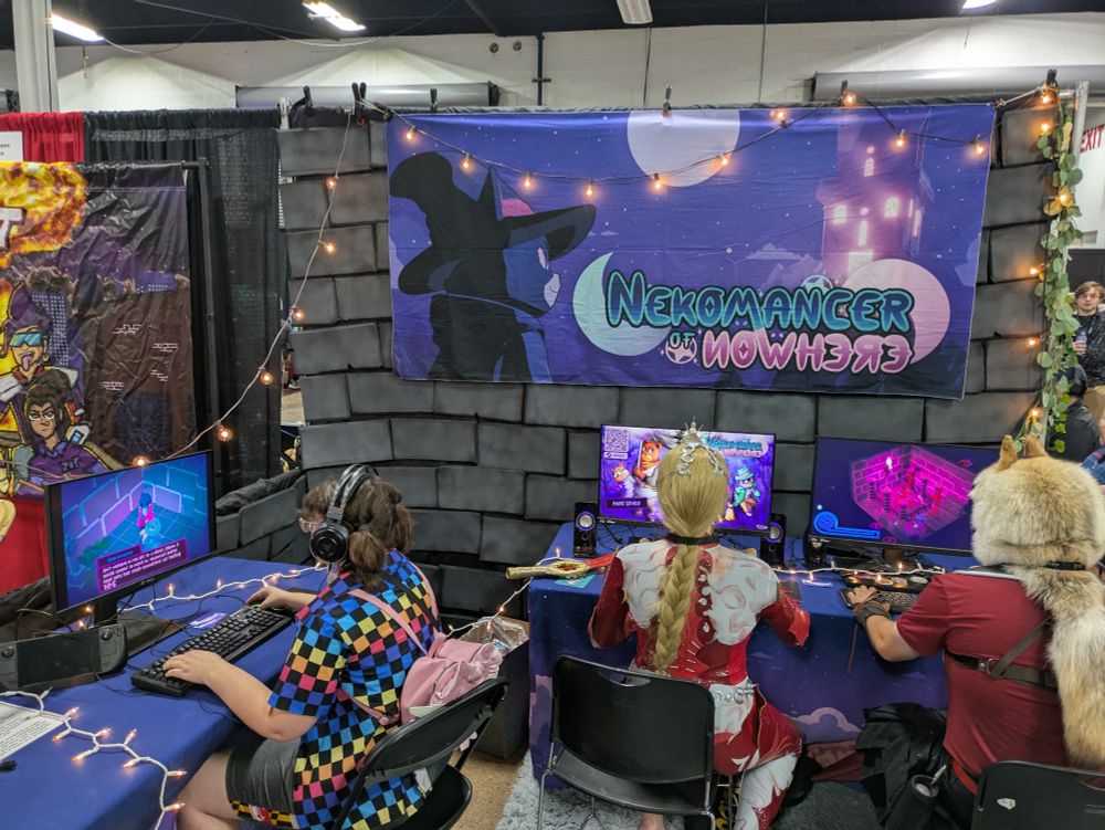 The Nekomancer of Nowhere booth! A large banger hangs on a brick wall, with lights around it. Three computers are playing the game
