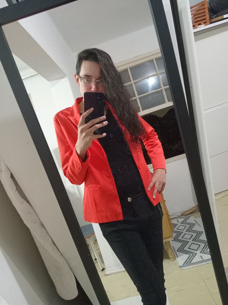 Amanda em frente a um espelho, ela está com uma calça jeans preta, uma blusa preta de manga longa com um pequeno decote e um blazer laranja, seu cabelo está dividido para a direita, ela segura o celular com a mão esquerda com a mão direita na cintura