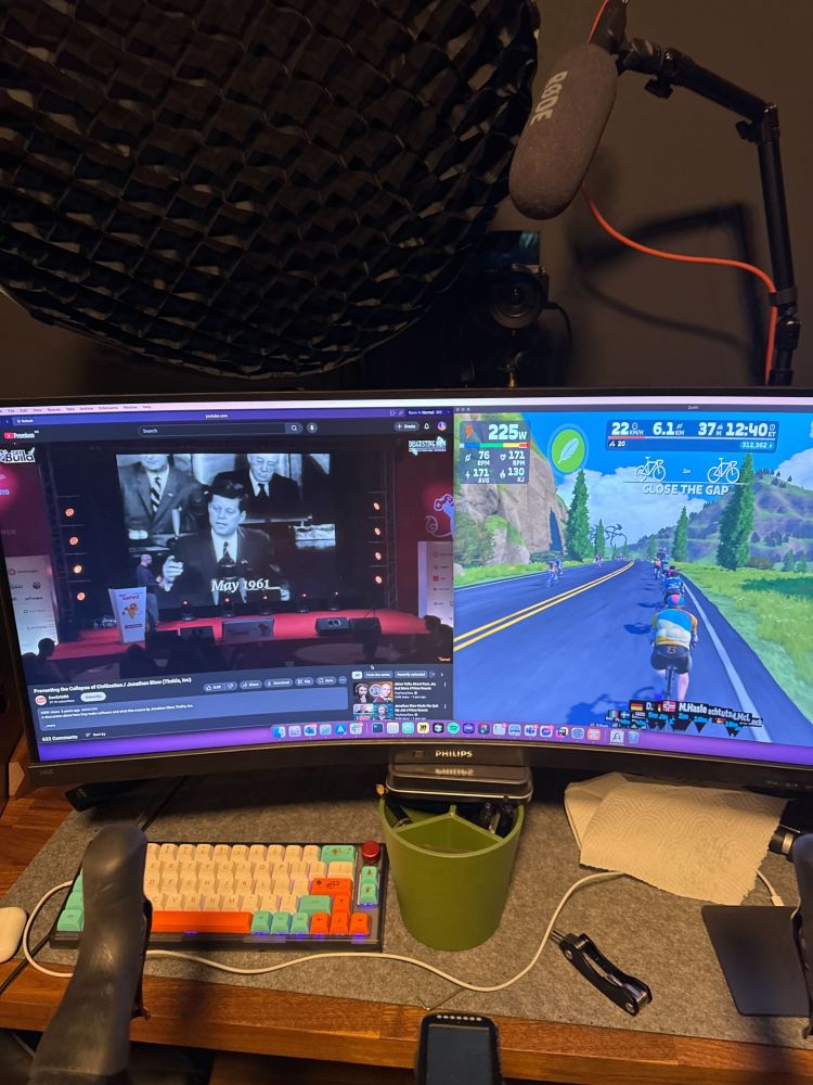 Bilde av en PC skjerm hvor du kan se en forelesning på den ene halvdelen og en sykkel-simulator på den andre halvdelen. Du kan også skimte elementer av en treningsykkel i nedre del av bildet og podcasting utstyr i øvre del av bildet.