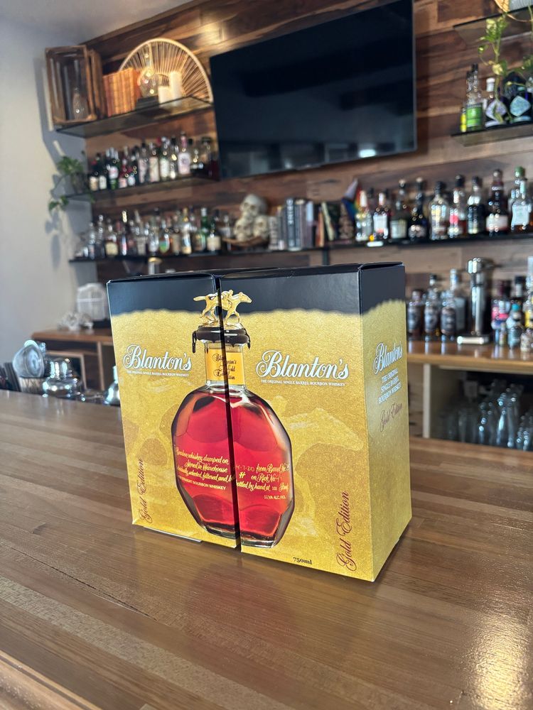 Two boxes containing bottles of Blanton’s Bourbon Gold Edition sit on a butcher block wooden bar counter in front of a nice bar. Blanton’s Gold is a very sought-after bottle.